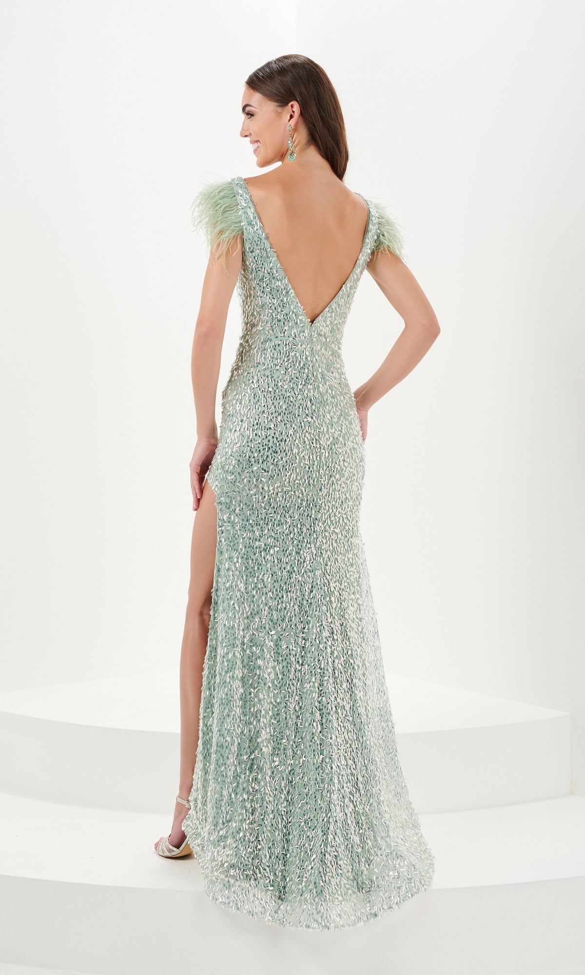 A woman wearing the Long Prom Dress 16055 by Tiffany—a light green sequin gown with feather-trimmed shoulders, deep V-back, and thigh-high slit—stands on white steps in high-heeled sandals, facing away.